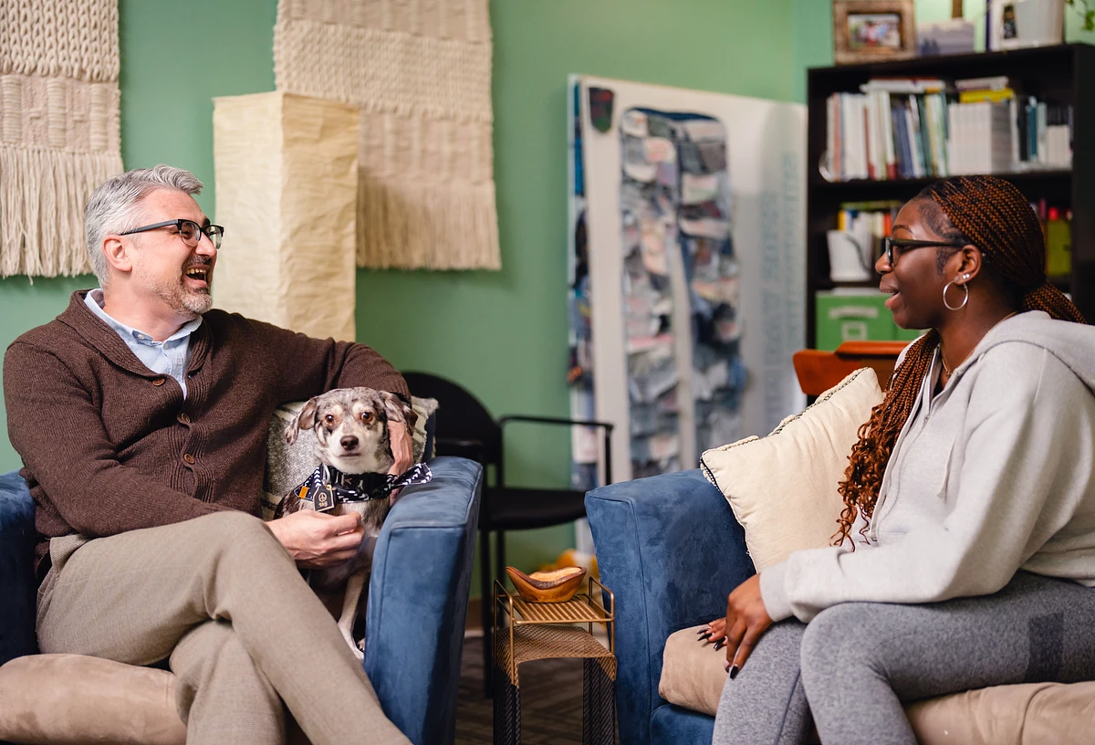 A man and a woman are sitting and conversing on chairs in a cozy room with a dog sitting on the man's lap.