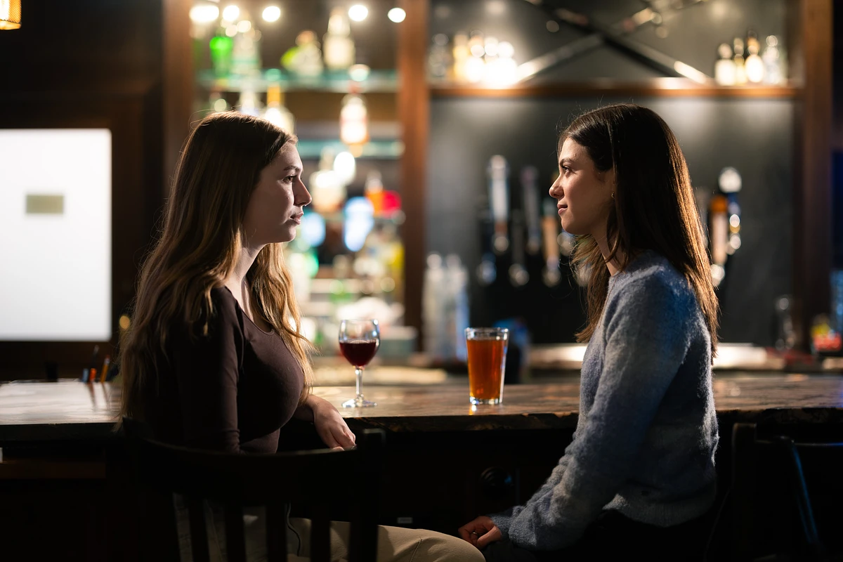 Two women are sitting at a bar, engaged in conversation with drinks in front of them.