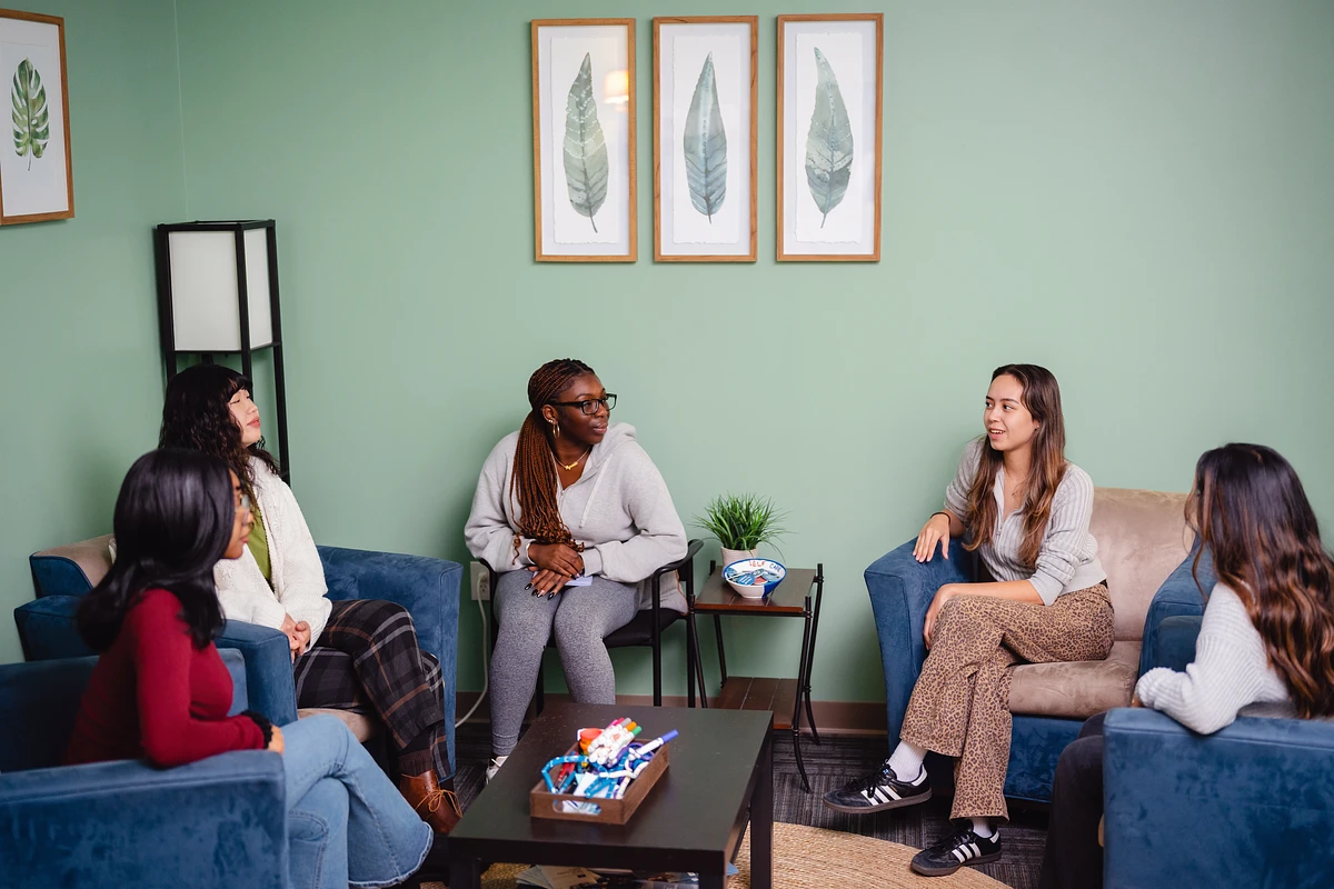A group of people are engaged in a discussion while sitting in a room with green walls and leafy artwork.